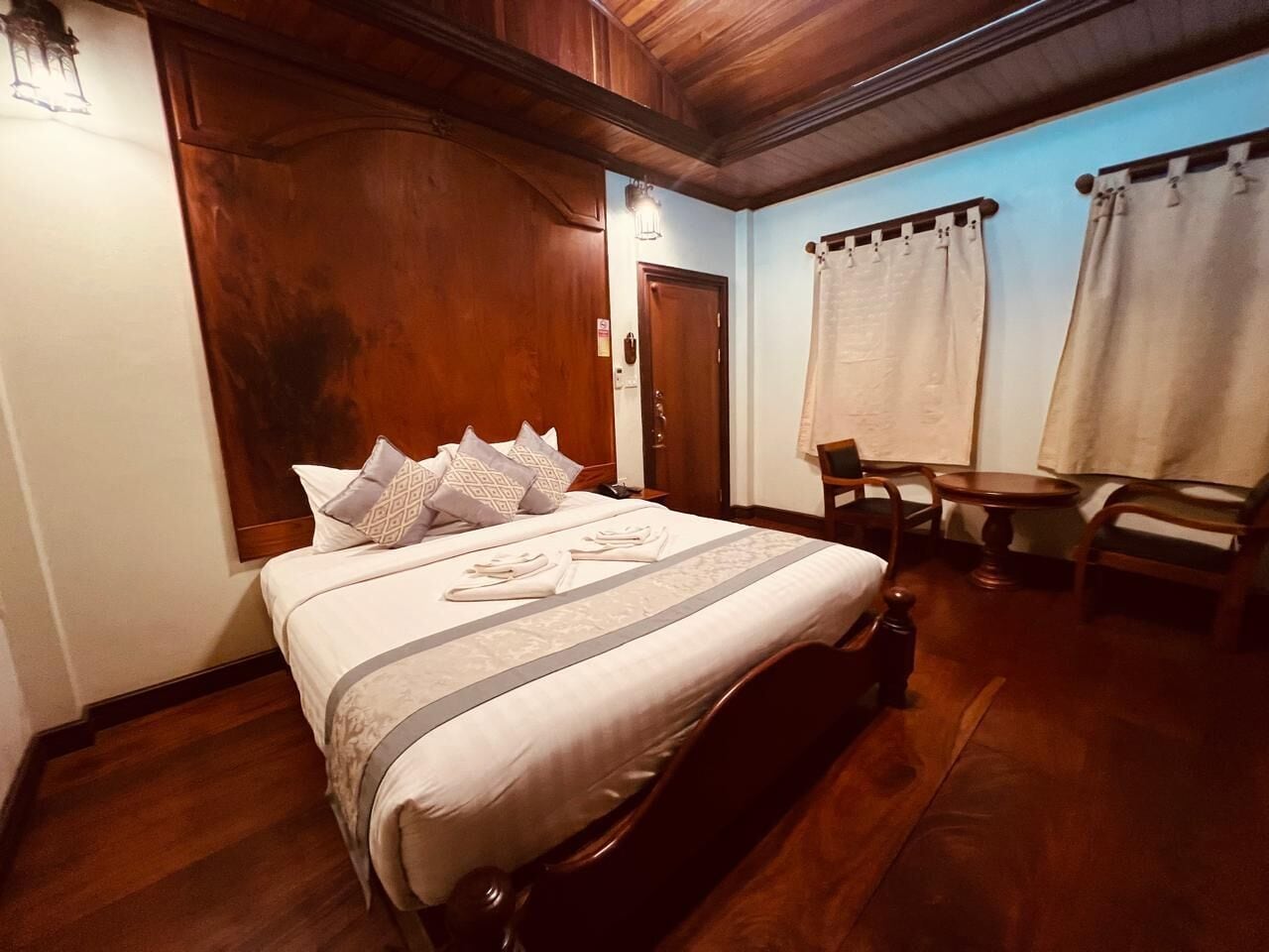A neatly made bed with a patterned bedspread and pillows, a wooden headboard, a small wooden table, and a chair in a room with wooden flooring and walls.