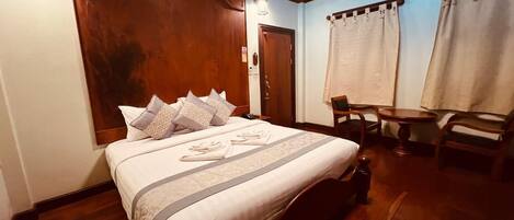 A neatly made bed with a patterned bedspread and pillows, a wooden headboard, a small wooden table, and a chair in a room with wooden flooring and walls.