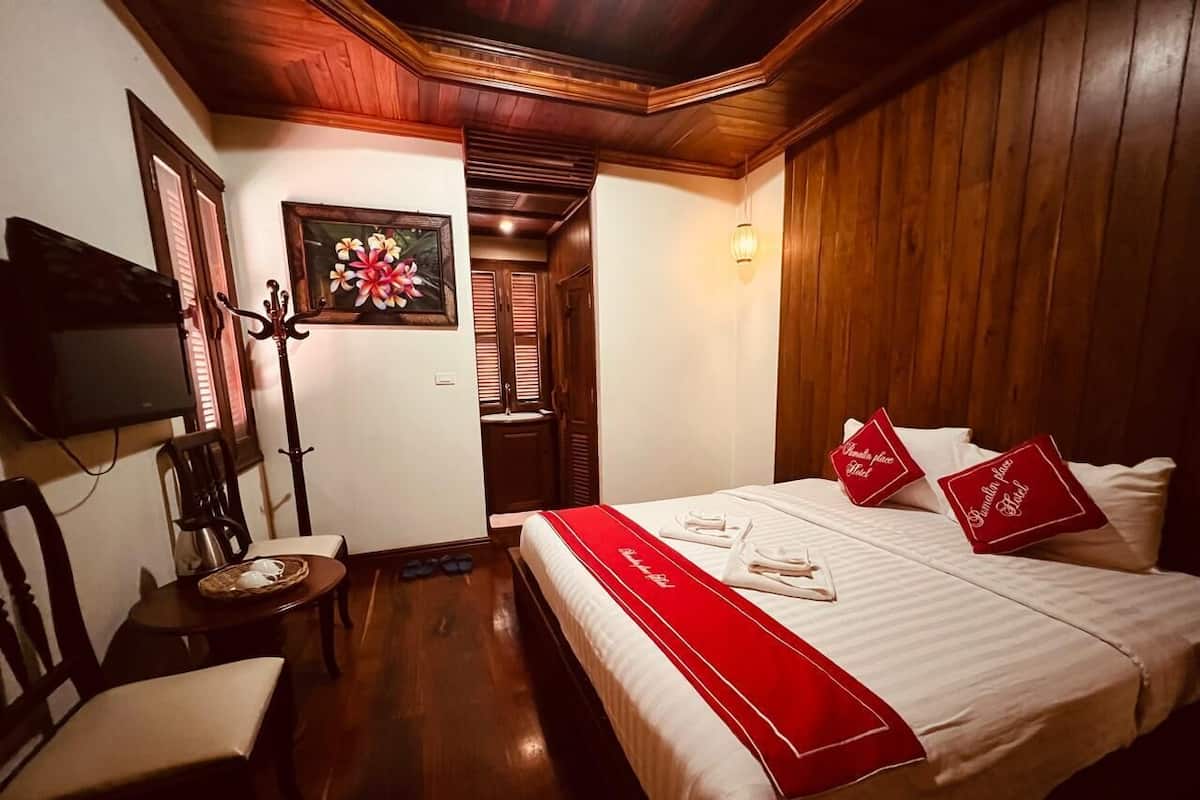 A hotel room with a wooden ceiling, a bed with red pillows, a red bed runner, a chair, a small table with a basket, and a wall-mounted TV.