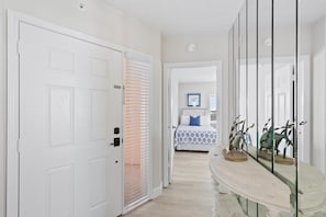 Corredor que dá para um quarto, com uma cama, mesa de cabeceira com objetos decorativos e uma porta com janela.
