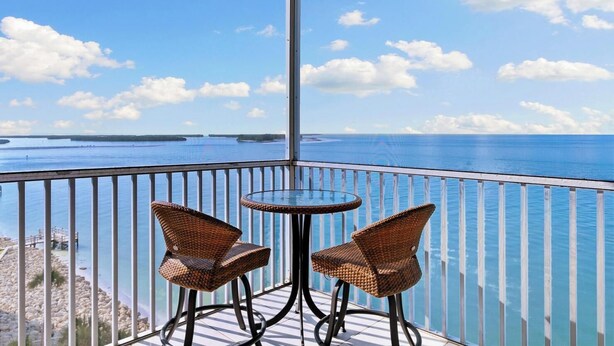 Ein Balkon mit zwei Korbstühlen und einem kleinen runden Tisch, mit Blick auf das klare, blaue Meer und entfernte Inseln.