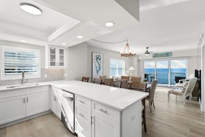A modern kitchen with white cabinetry, a large island, and a dining area with a view of the ocean. - Dela Park Place - Waterfront 2BDR/2BA with Den (Marco Island)