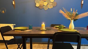 Un comedor con una mesa dispuesta para dos personas, con una vela, platos verdes y un jarrón con plantas secas.