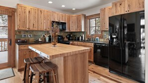 A modern kitchen with wooden cabinets, a central island with stools, a black refrigerator, and a window providing natural light.