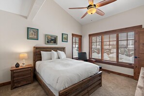 Un dormitorio con cama de madera, mesita de noche con lámpara, ventilador de techo y ventana con persianas.