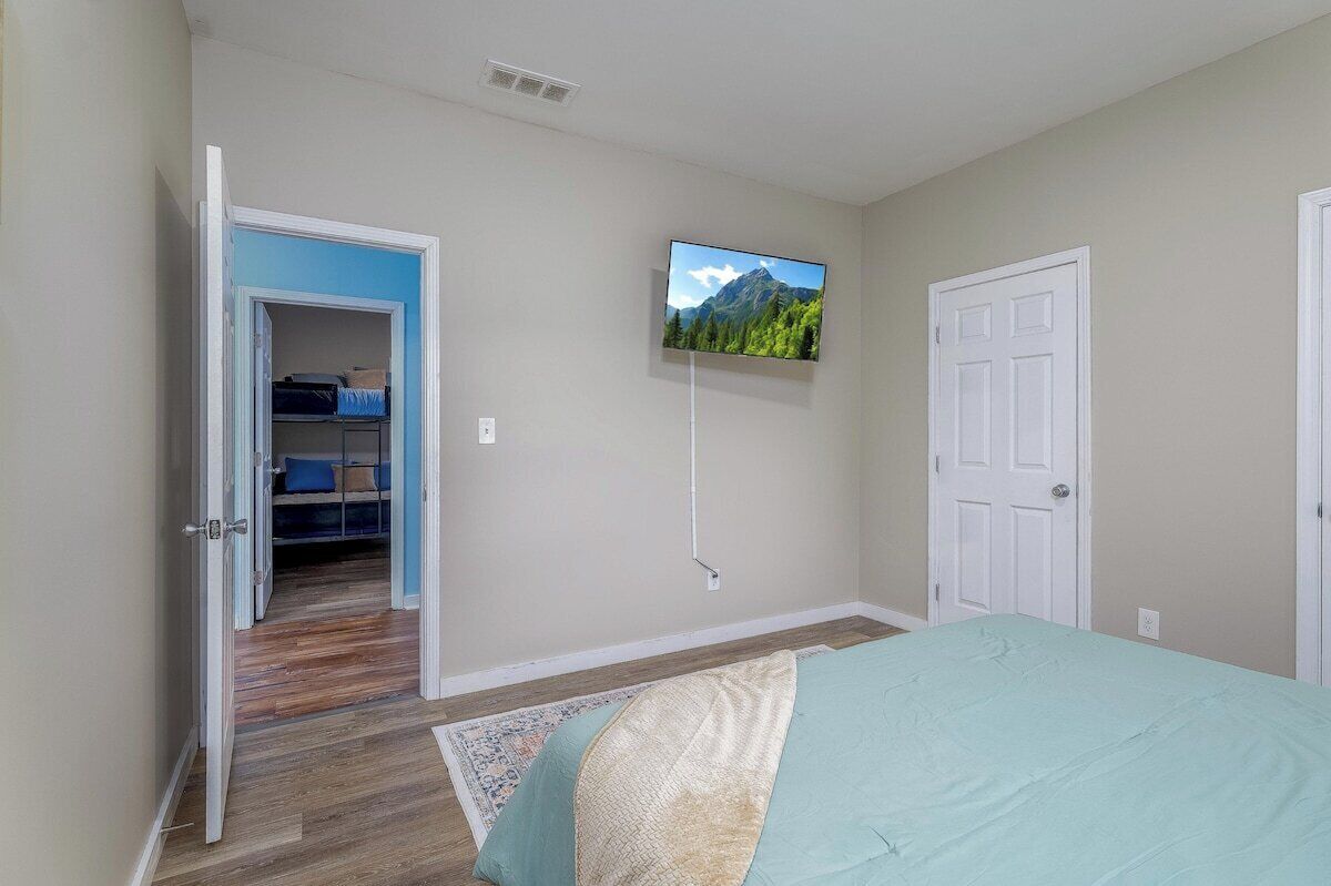 A bedroom with a bed, a TV mounted on the wall, a door, and a hallway leading to a closet.