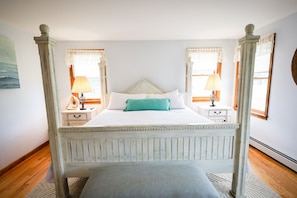 A four-poster bed with a white headboard, two bedside tables with lamps, a bench at the foot of the bed, and a painting on the wall.