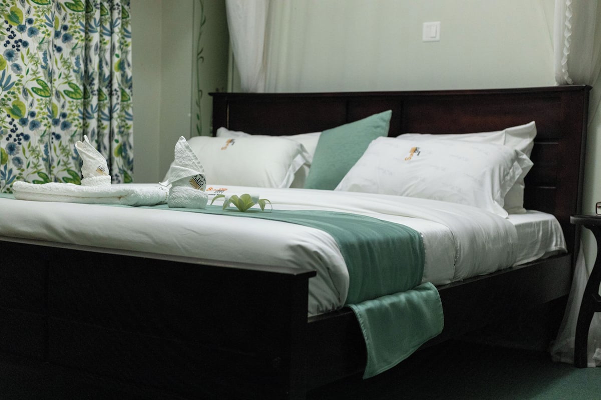 Un lit bien fait, avec du linge de lit blanc et un plaid vert, une tête de lit en bois et, en arrière-plan, un rideau à motifs floraux.
