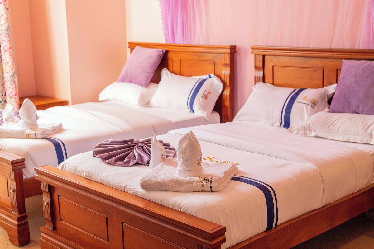 Deux lits doubles avec des têtes de lit en bois et du linge de lit blanc, chacun agrémenté d’un coussin violet et d’une serviette pliée posée sur le lit.