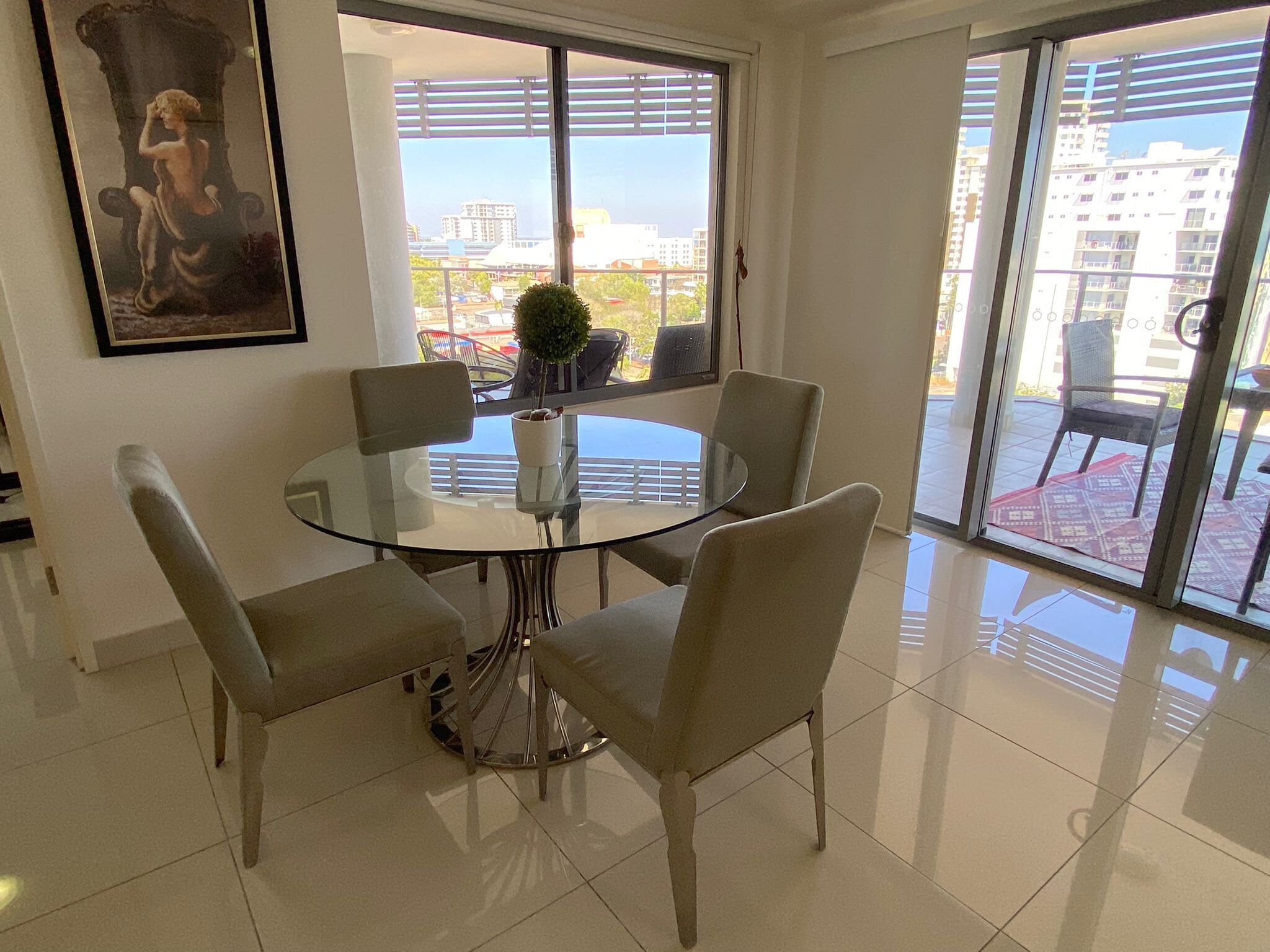 A dining area with a glass table and four chairs, a potted plant, and a painting on the wall.