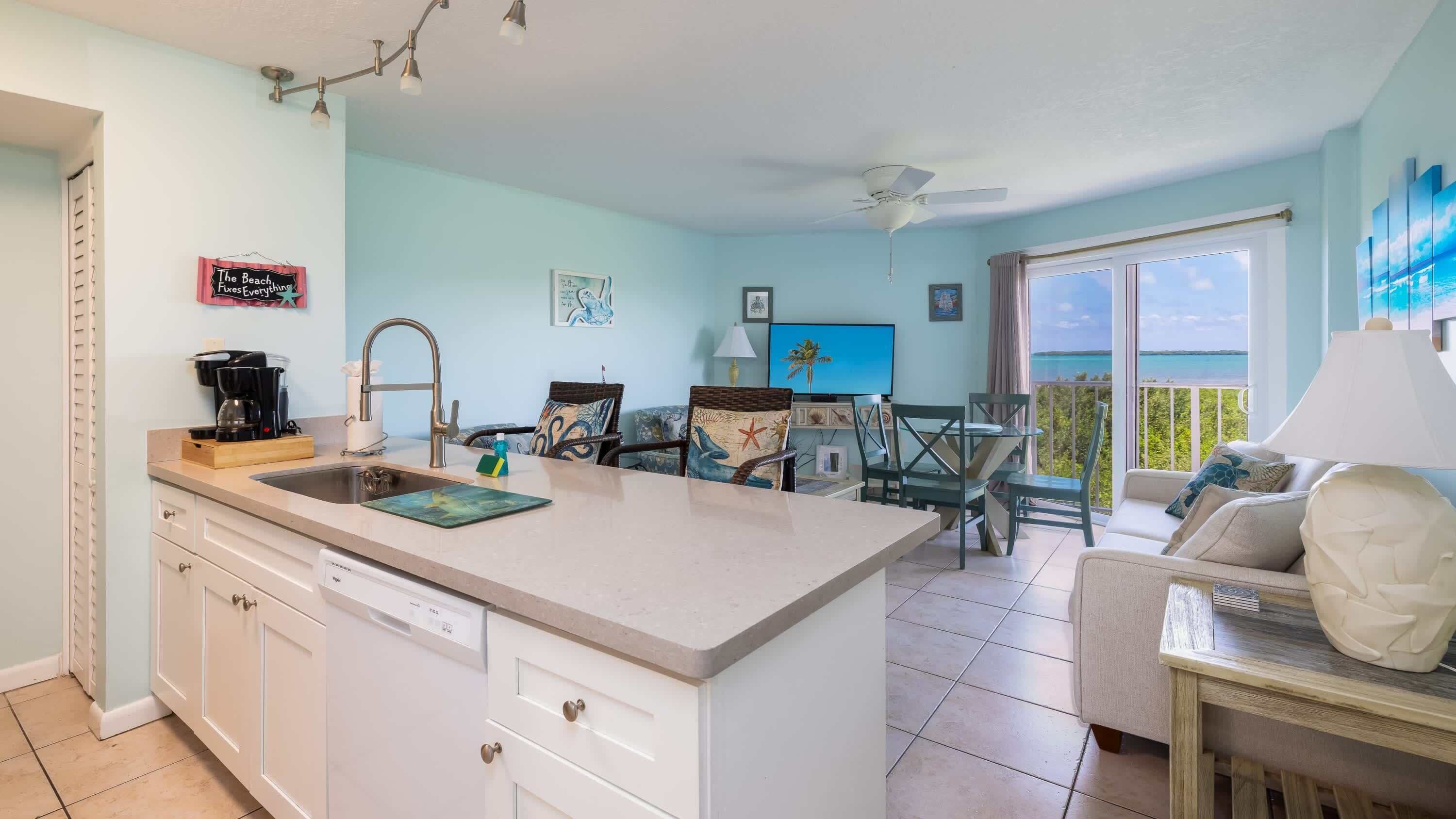 A kitchen with a central island, a dining area with chairs, a sofa, and a view of the sea.
