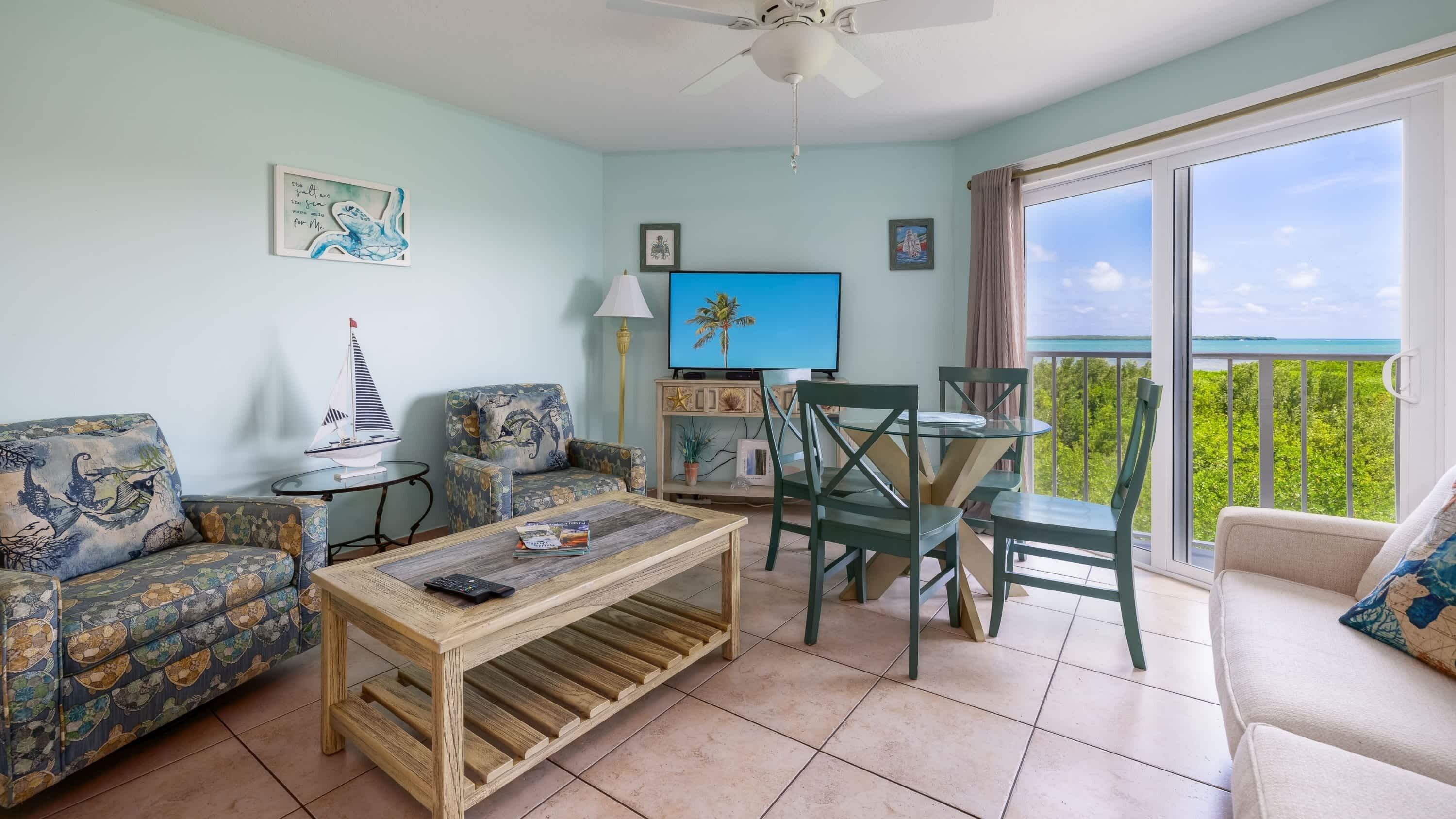 A living room with a sofa, armchair, coffee table, and a dining area with chairs and a table. There is a TV on the wall and a balcony with a view.