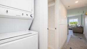 A laundry room with a FRIGIDAIRE washer and dryer, a door with louvered shutters, and a view into a living area with a sofa and a window.