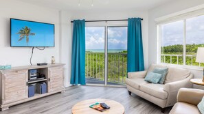 A room with a sofa, a coffee table, a TV, and a balcony with curtains. - Nauti Hideaway | 2 Balconies | Ocean Pointe 2314 (Tavernier)