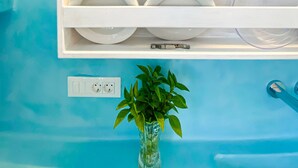 Un bagno con uno scaffale bianco che contiene stoviglie e bicchieri bianchi, un vaso trasparente con piante verdi e una parete blu.