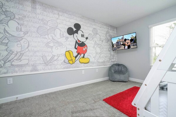 A room with a Mickey Mouse mural, a television, a gray bean bag chair, and a red rug.