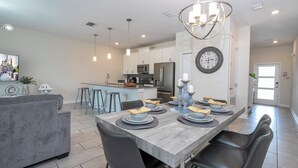 A modern kitchen with white cabinets, stainless steel appliances, and a dining area with a grey table set for four. - Modern 4Bed+PrivatePool Resort Style 8959 (Davenport)
