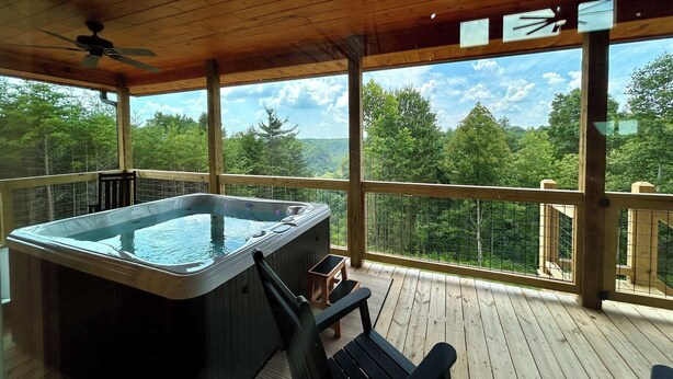 A wooden deck with a hot tub, chairs, and a view of a forested landscape. - Maple Moon | Hot tub | WiFi | New | Views (Rogers)