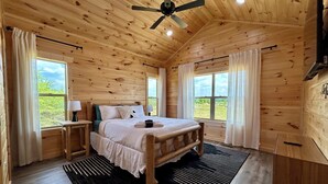 A wooden cabin bedroom with a bed, nightstands, a ceiling fan, and large windows. - Maple Moon | Hot tub | WiFi | New | Views (Rogers)