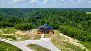 A cabin with a sloped roof, surrounded by dense forest, with a gravel driveway leading to the entrance. - Maple Moon | Hot tub | WiFi | New | Views (Rogers)