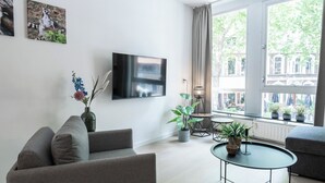 Ett modernt vardagsrum med en grå soffa, en platt-TV, ett litet runt bord med en växt och utsikt över byggnader genom stora fönster.