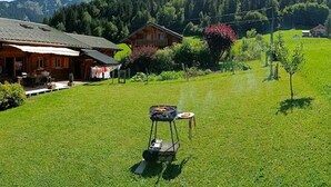 A wooden cabin with a porch, a barbecue grill, and a grassy field with trees and mountains in the background. - Spacious chalet with WIFI, pets allowed, private garage (Abondance)