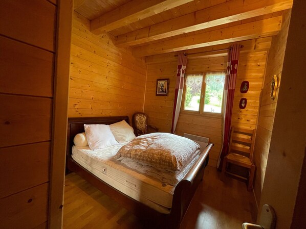A wooden cabin bedroom with a bed, a chair, a ladder, and a window with curtains.