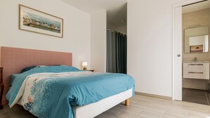 Een slaapkamer met een bed, een nachtkastje, een lamp, een schilderij aan de muur en een zichtbare badkamer door een open deur.