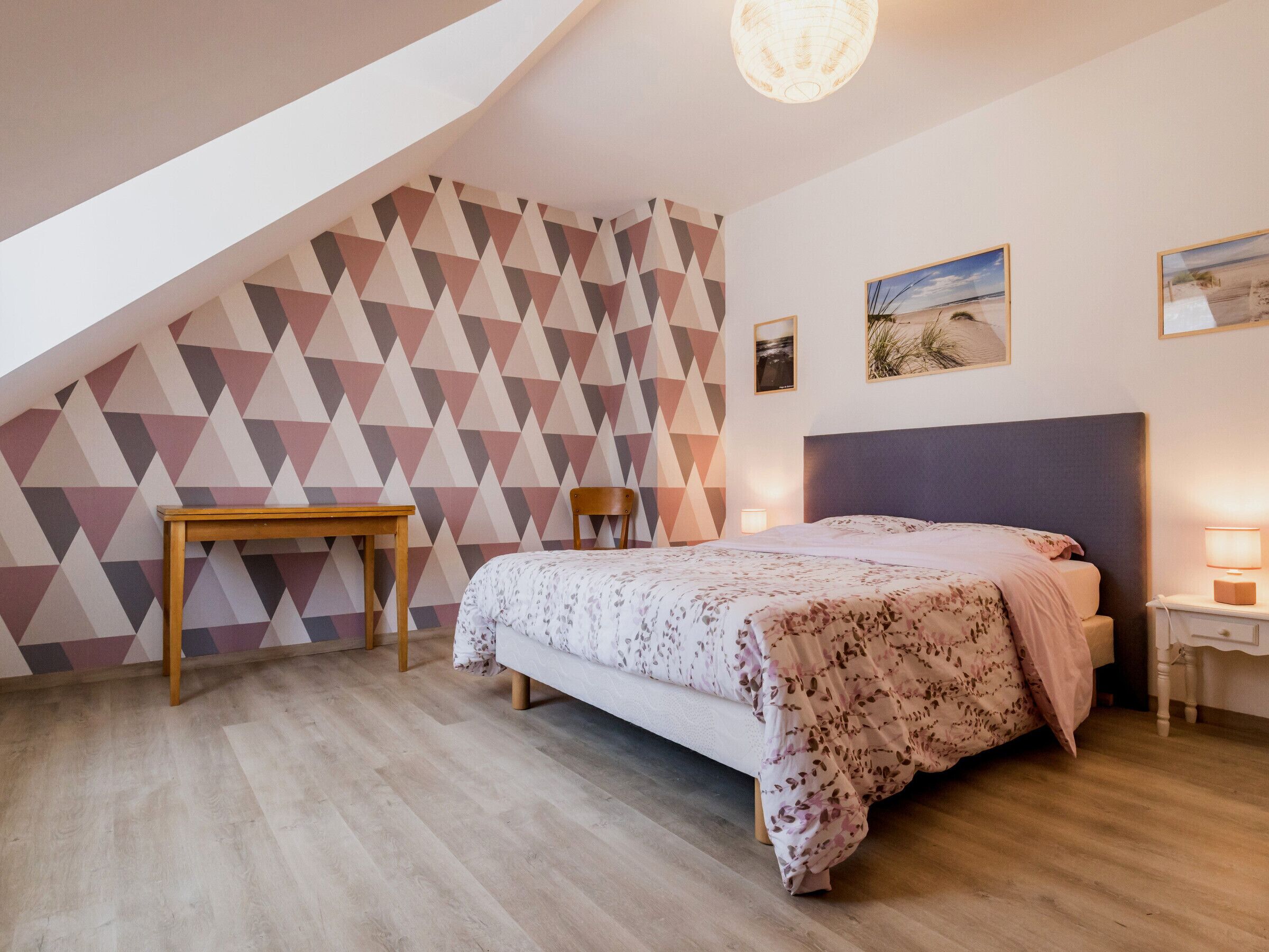 Une chambre avec un plafond en pente, un lit recouvert d’une couvre-lit à motifs floraux, une table de chevet en bois et un mur orné d’un motif géométrique.