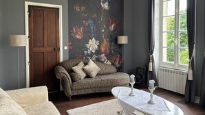A living room with a dark grey wall, a floral mural, a brown door, a patterned sofa, a white table with candles, and a large window with curtains.