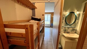 Habitación pequeña con literas, un escritorio, un baño con lavamanos y espejo, y vista a una ventana y una puerta que da a otra habitación.