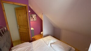 A bedroom with a bed, a wooden door, a small bathroom, and a sloped ceiling. - Charming 50m² Chalet Apartment - WiFi, Ski Storage, Near Slopes & Shuttle (Le Lioran)