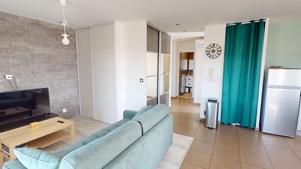 A modern living room with a sofa, a TV, and a wooden coffee table. There is a kitchen area with a fridge and a teal curtain. - Quiet Apartment with Terrace, Parking, and AC, 5 Min Drive to Beach (Frontignan)