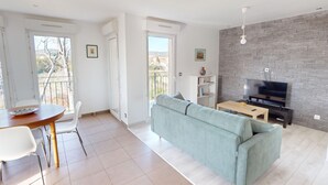 A modern living room with a sofa, a dining table with chairs, a TV, and a bookshelf. There are large windows and a balcony. - Quiet Apartment with Terrace, Parking, and AC, 5 Min Drive to Beach (Frontignan)