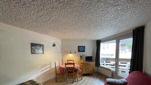 A compact room with a bed, a desk with a chair, a dining table with chairs, and a large window. - Ski-in/Ski-out Studio, South-East Balcony, Sleeps 4, Shops Onsite (Le Lioran)