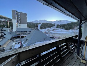 A view from a balcony overlooking a snowy landscape with mountains, a ski slope, and a building with multiple floors.