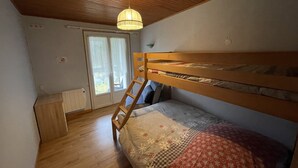 A small, cozy room with a bunk bed, wooden flooring, a ladder, a window with curtains, and a ceiling light.
