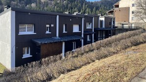 A row of modern, dark-colored buildings with multiple windows and balconies, set against a backdrop of a forested hillside.