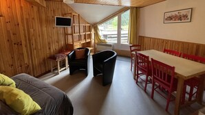 A cozy room with wooden walls and ceiling, a sofa, a dining table with chairs, a TV mounted on the wall, and a window with curtains. - Central Duplex with Balcony – Sleeps 6/8, Pets Welcome (Le Lioran)