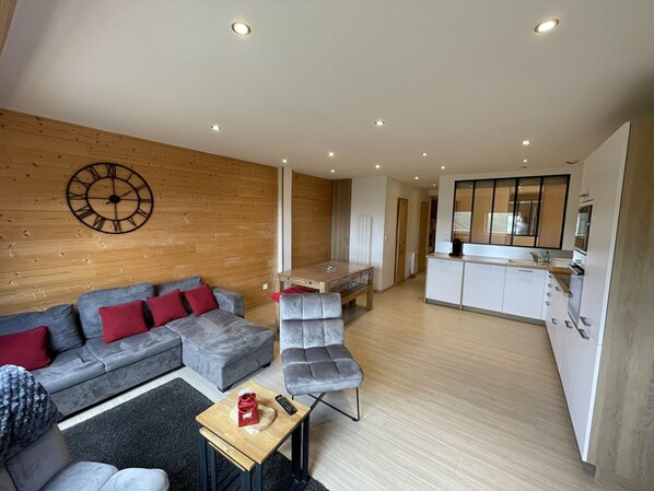 Un salon moderne avec un canapé d’angle gris, une horloge murale en bois et une petite table en bois sur laquelle repose une télécommande.