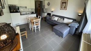 A modern living room with a grey sofa, a wooden dining table with chairs, a kitchen area with white cabinets, and a clock on the wall. - Central studio with balcony and WiFi (Le Lioran)
