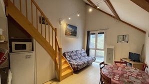 A cozy living space with a wooden staircase, a sofa, a dining table, and a kitchen area with a microwave. - Ski-In/Ski-Out Studio with Mezzanine, Balcony & Free Shuttle (Le Lioran)