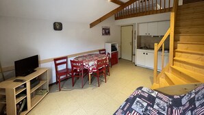 A cozy living room with a wooden staircase, a dining area with a table and chairs, and a kitchen with white cabinets. - Ski-In/Ski-Out Studio with Mezzanine, Balcony & Free Shuttle (Le Lioran)