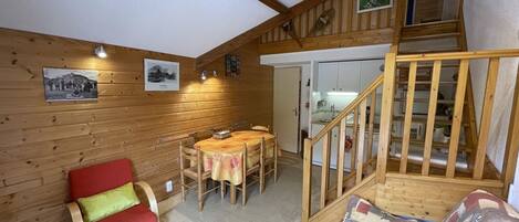 A cozy cabin interior with a dining area, a red sofa, a wooden staircase, and a kitchen visible through an open door.