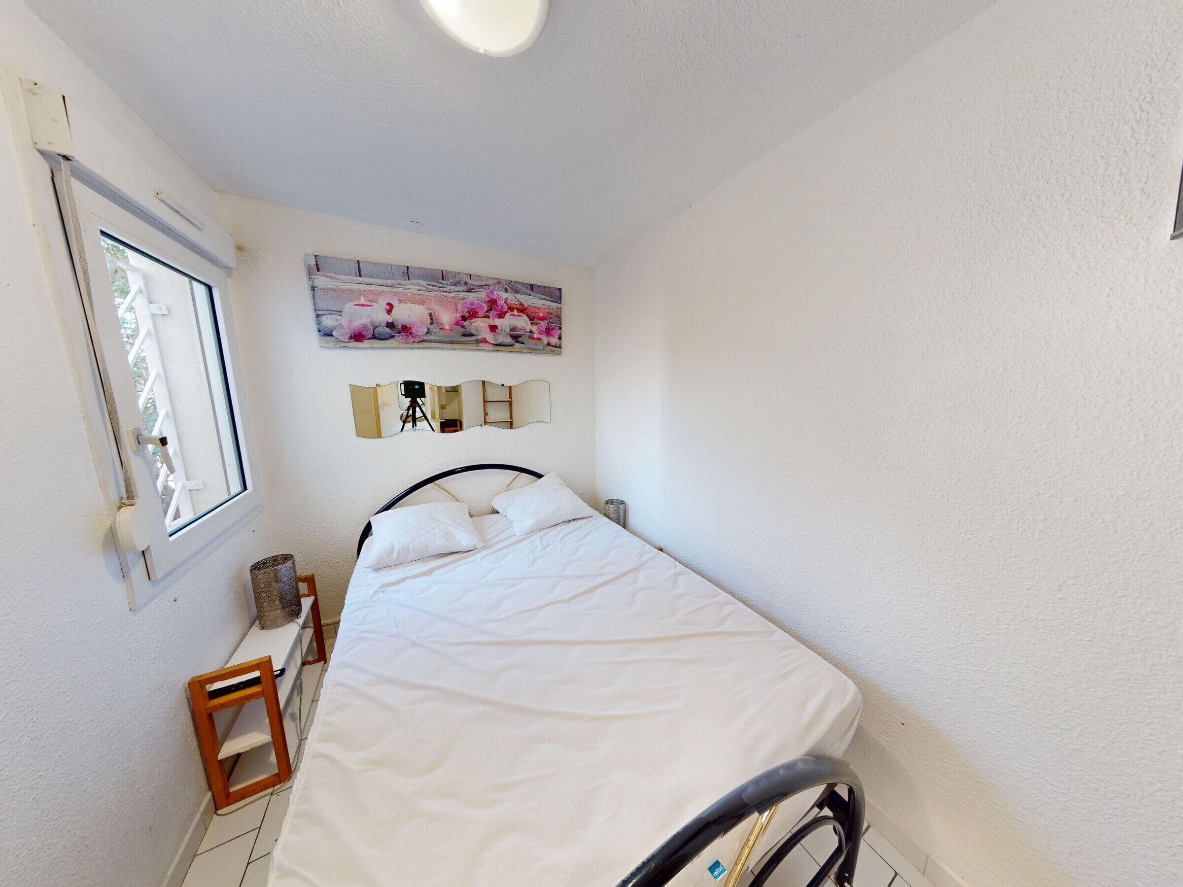 A bedroom with a bed, a window, a small stool, a mirror, and a painting on the wall.