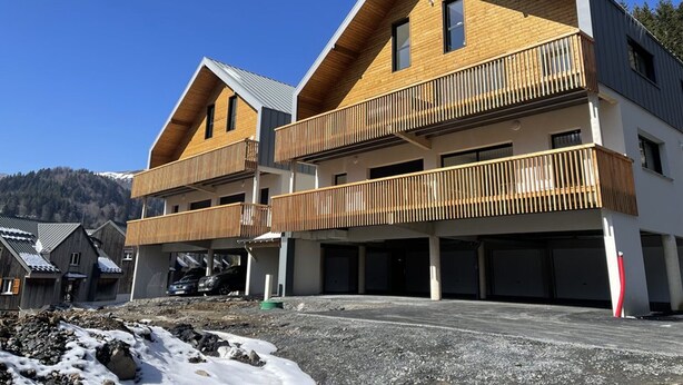 A modern multi-story building with wooden balconies, a garage area, and a mountainous backdrop. - Cozy New 2-BR Apartment with Large Balcony, Close to Chairlift (Le Lioran)