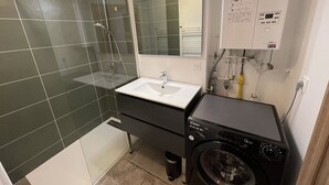 A modern bathroom with a glass-enclosed shower, a white sink, a black washing machine, and a mirror. - Cozy New 2-BR Apartment with Large Balcony, Close to Chairlift (Le Lioran)