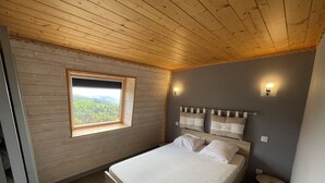A bedroom with a wooden ceiling, a window with a view of greenery, a bed with white bedding, and two wall-mounted lights. - Charming duplex near slopes with balcony and parking (Saint-Jacques-des-Blats)