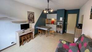 A compact living space with a kitchenette, a dining area with stools, a sofa, and a TV. - Charming Studio Facing Ski Slopes - Pet Friendly, South Exposure (Le Lioran)