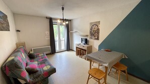A cozy living room with a sofa, a dining table with chairs, a TV, and a staircase. - Charming Studio Facing Ski Slopes - Pet Friendly, South Exposure (Le Lioran)